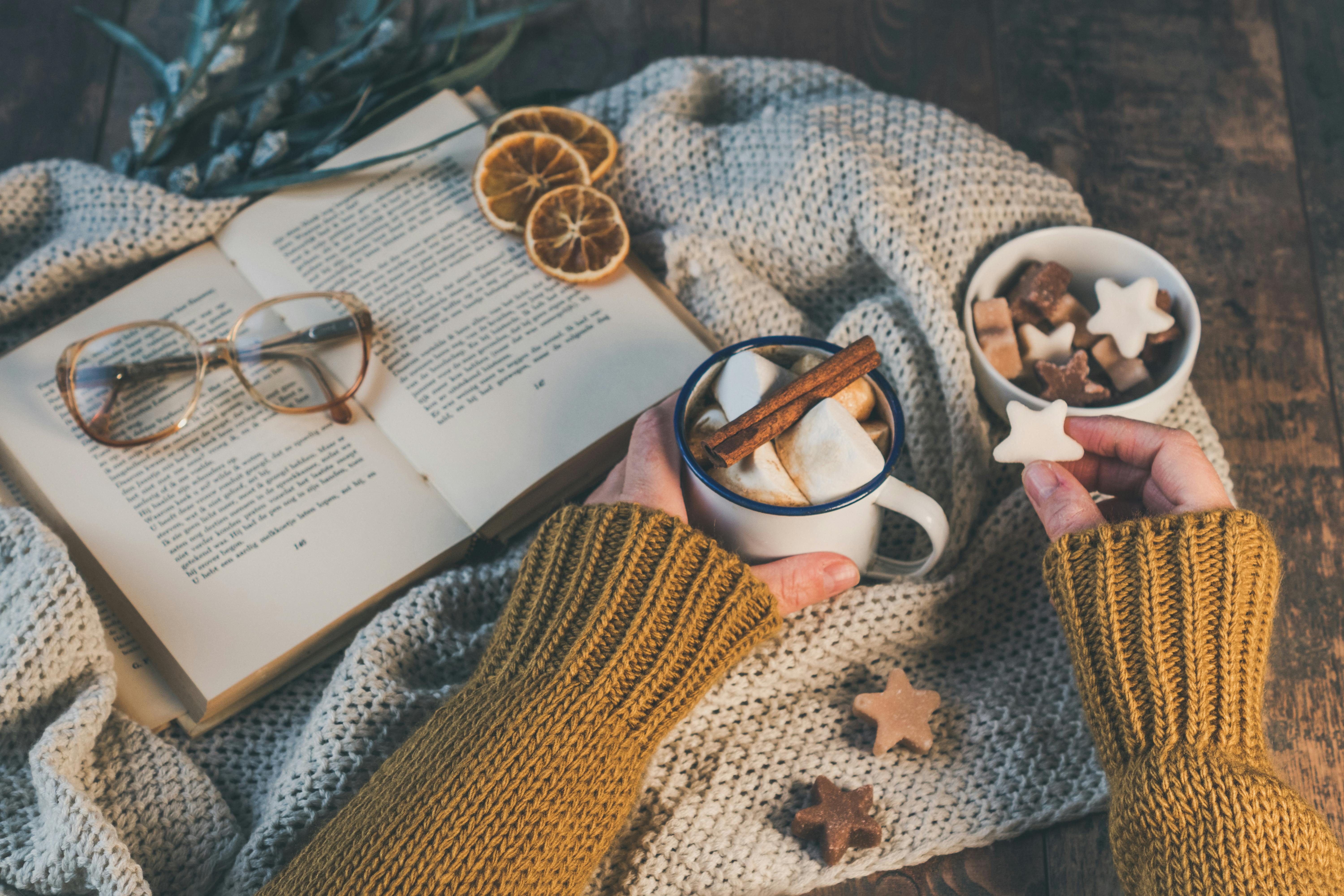 Woman's hands holding a mug of hot chocolate and a book on a wooden table, evoking a cozy fall atmosphere.