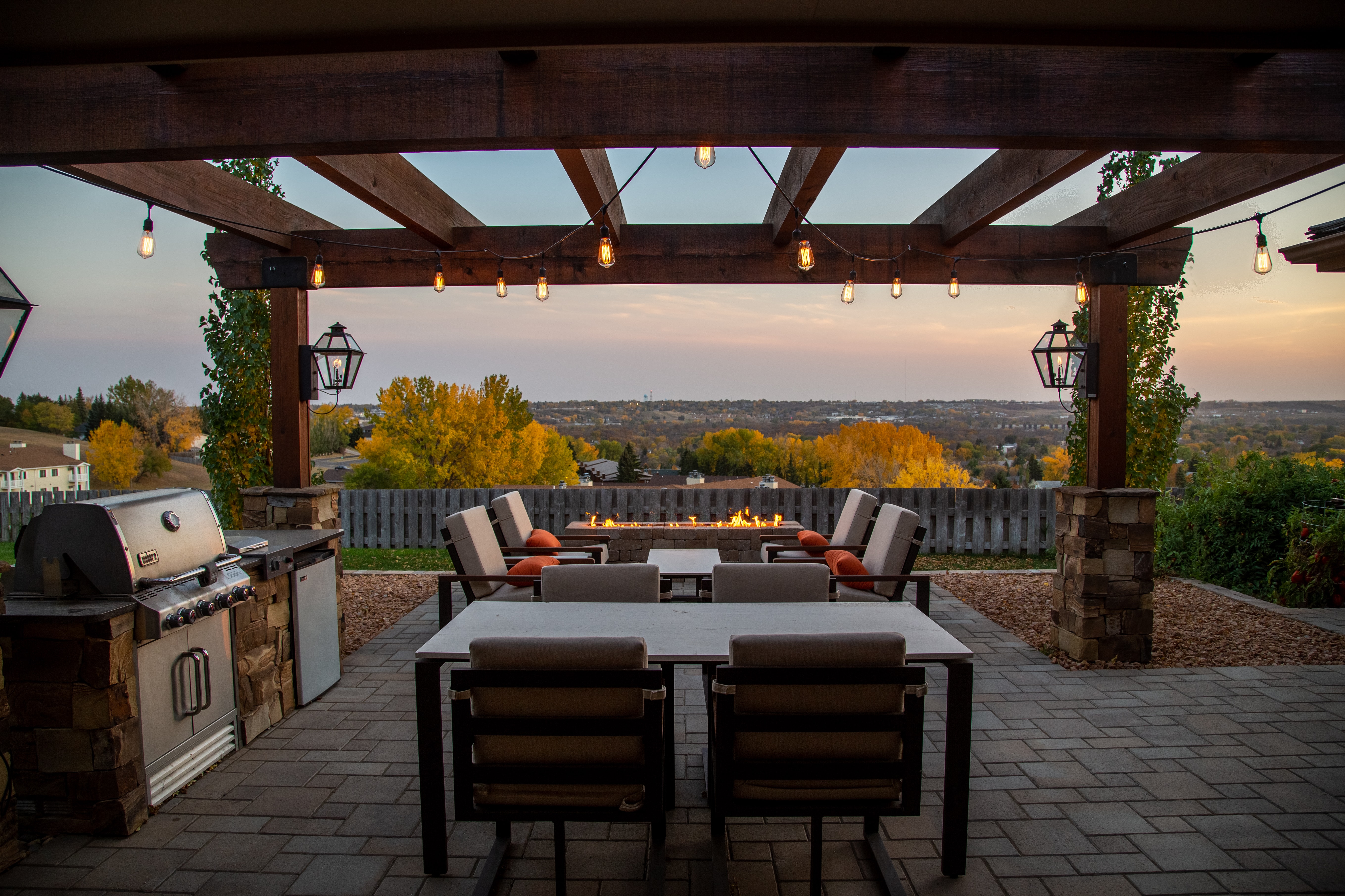 View of patio in residential yard. Pergola above outdoor dining area with string lights; a firepit in the distance.